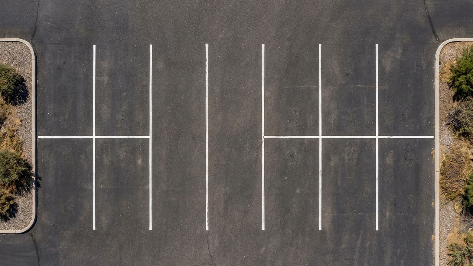 Painted parking stall lines on dark asphalt seen from directly above, a run of marked stalls divided by a continuous line down the middle, gravel and dry brush at both edges, no vehicles and no lettering anywhere